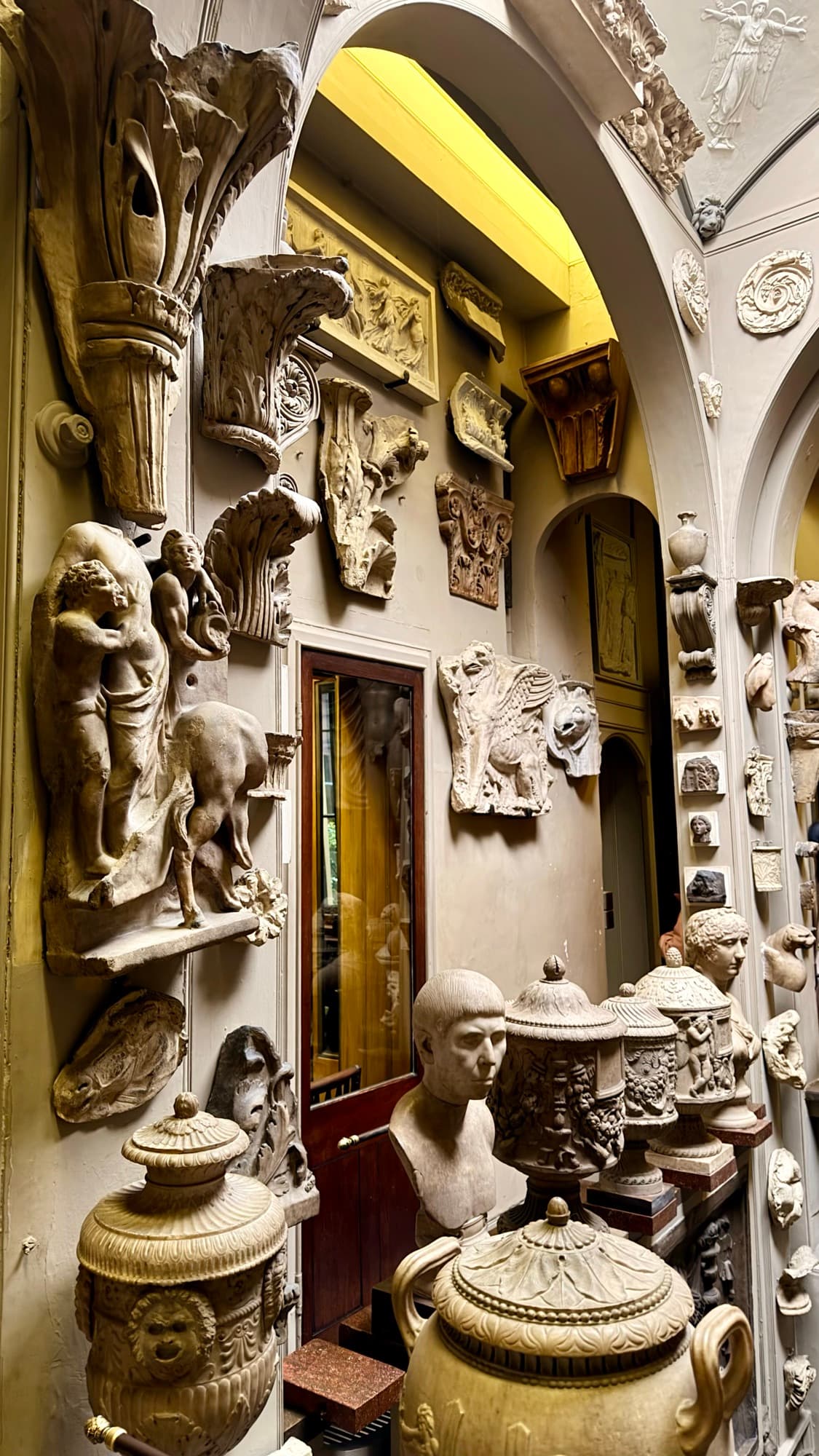 A museum corridor packed wall to ceiling with plaster casts, carved fragments and lidded funerary urns.