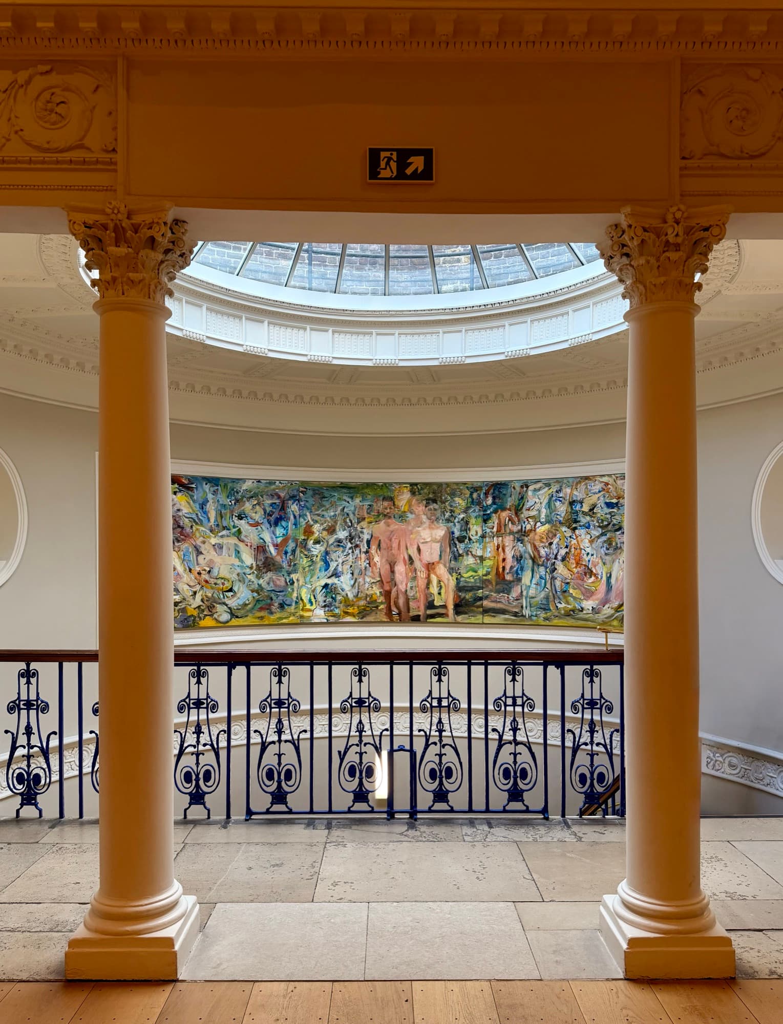 A gallery landing framed by two painted columns, a glazed dome above and a mural over the ironwork balustrade.