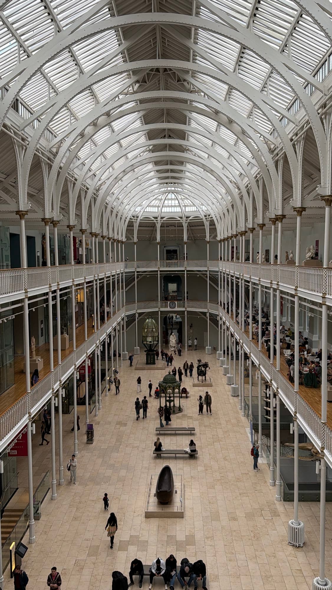 A museum's grand gallery seen from the upper balcony, white cast-iron arcades running under a barrel-vaulted glass roof.