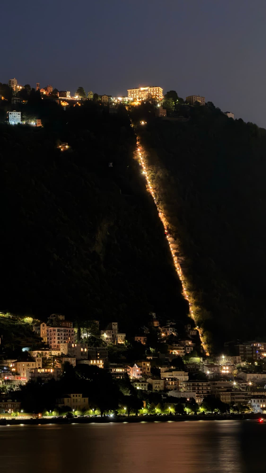 A funicular line lit in a single thread up a dark wooded hillside at night, above a lakeside town and the water below.
