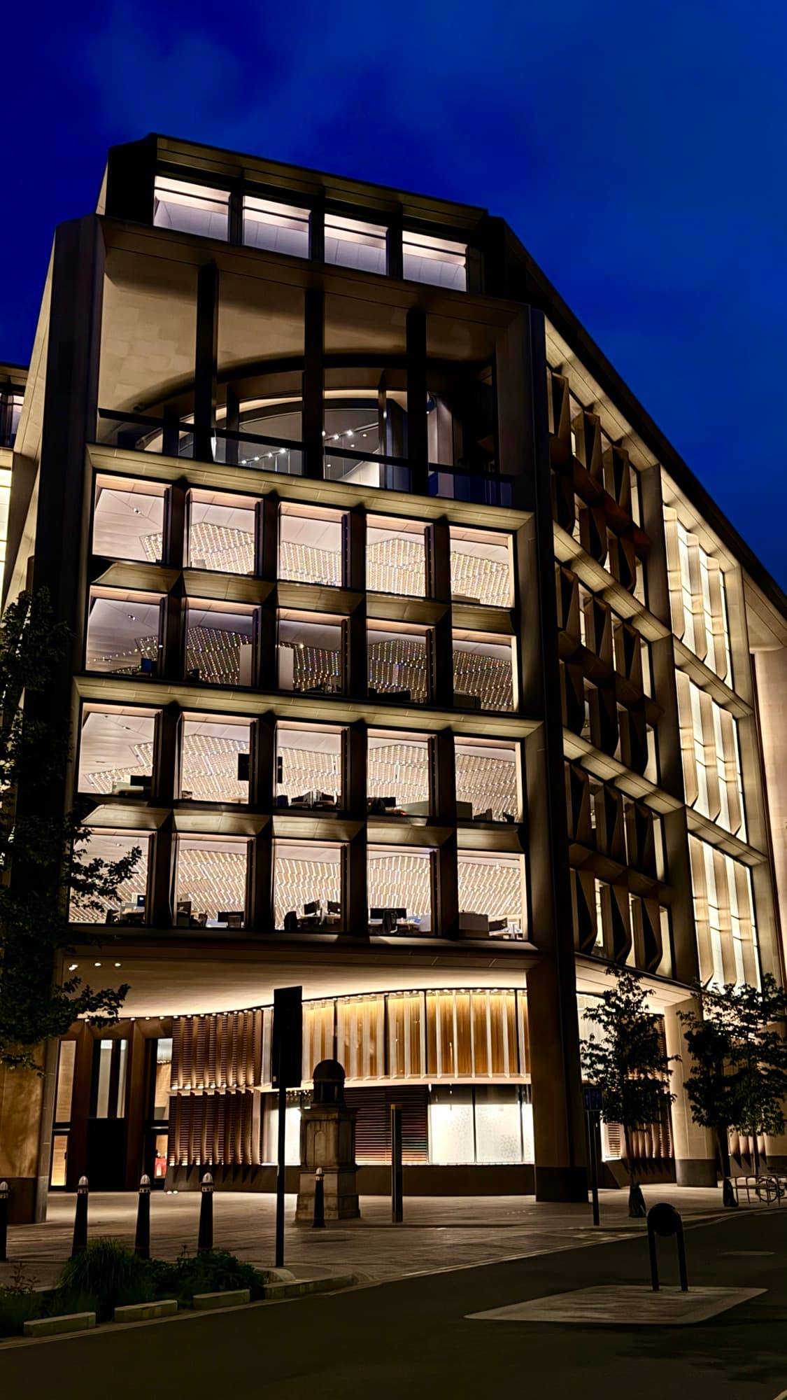A modern stone and glass office block lit from the inside at blue hour, empty street in front.