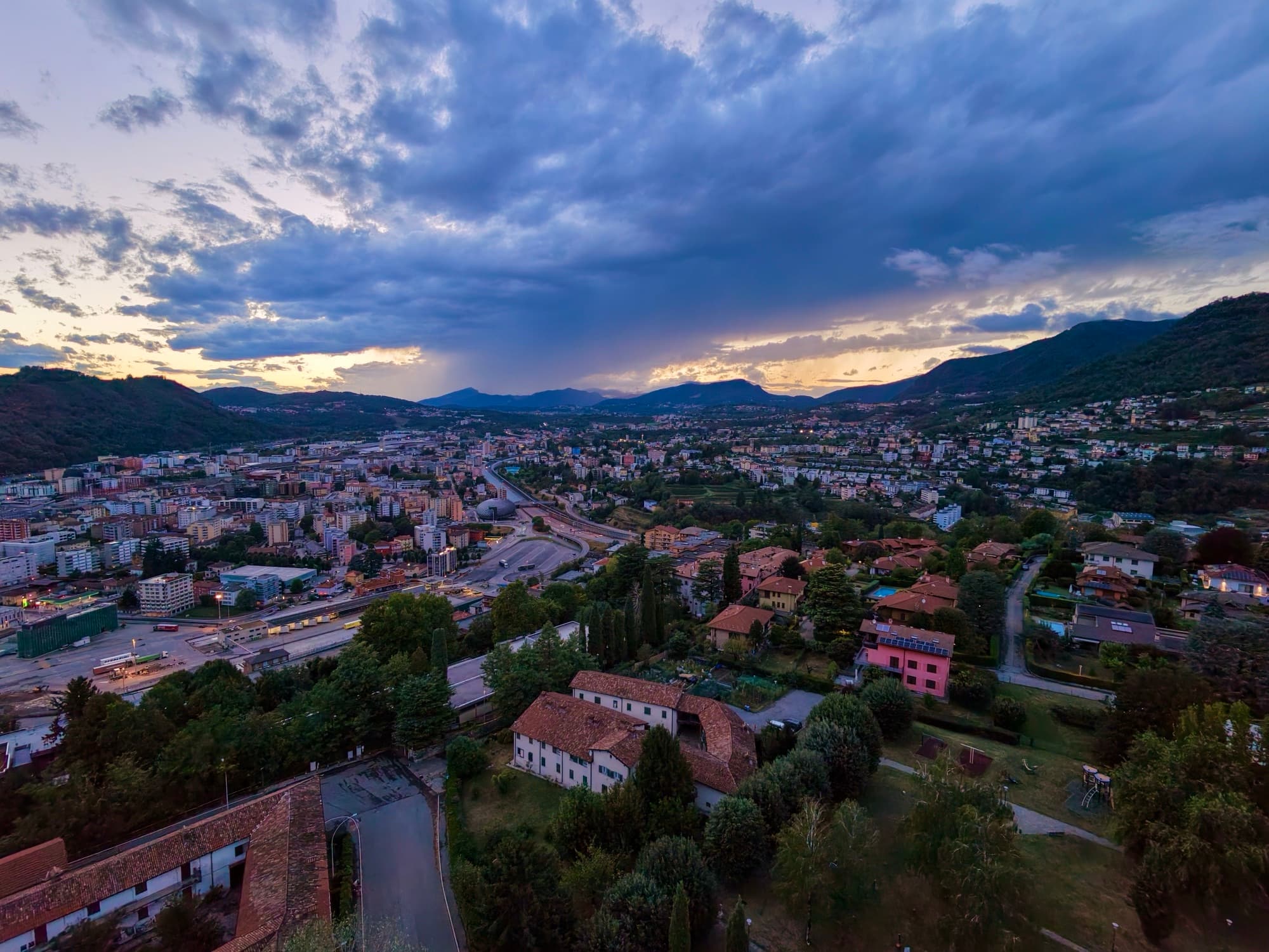 An aerial view over a valley town at dusk, a storm cloud breaking open above the ridgeline behind it.
