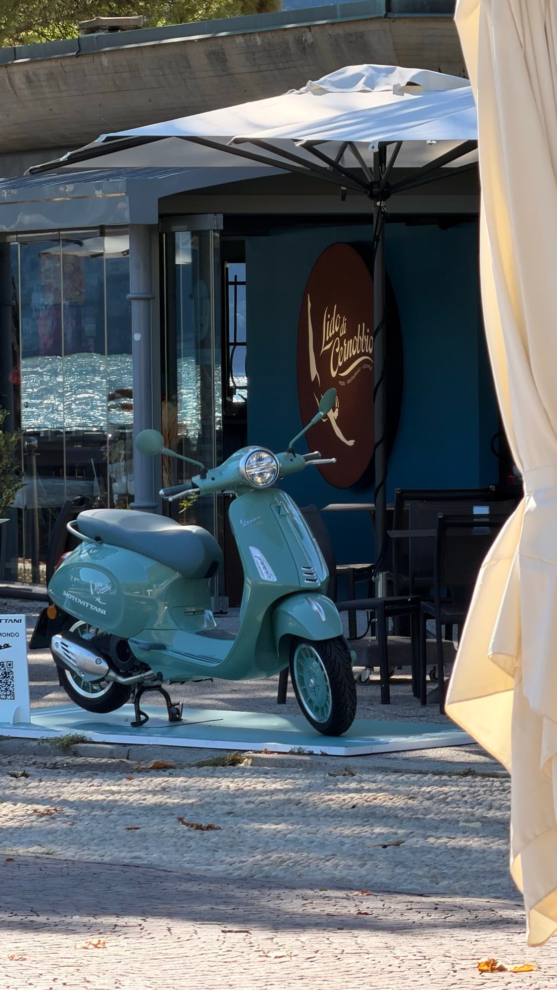 A sage green Vespa on a white display plinth outside a cafe, under a parasol.